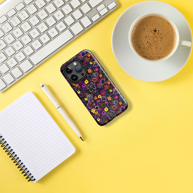 An iPhone, adorned with the vibrant In the Midnight Garden MagSafe case by My Favourite Colour is Rainbow, rests on a yellow surface. Nearby are a keyboard, coffee cup, notebook, and pen. The bright, orderly scene suggests an organized workspace.