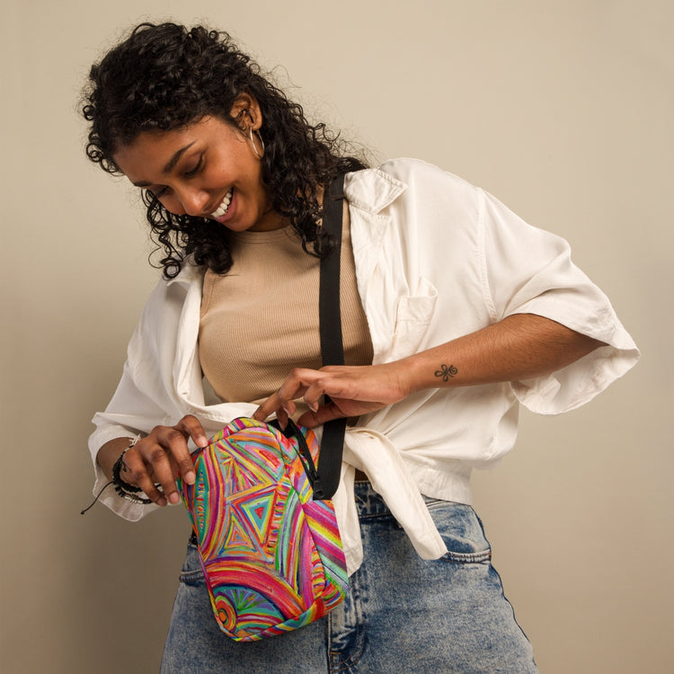 A person with curly hair, in a white shirt over a beige top and jeans, smiles as they open the Karma Kaleidoscope Mini Crossbody Bag by My Favourite Colour is Rainbow. Its vibrant print stands out against a neutral background, and adjustable straps rest comfortably on their shoulder.