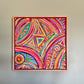 The Karma Kaleidoscope Framed Original Painting by My Store features vibrant swirls, triangles, and circles in pink, purple, orange, and yellow hues. Elegantly framed in simple wood, it is displayed on a plain light gray wall.