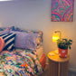 A colorful bedroom corner features a bed with vibrant, patterned bedding. A wooden side table supports a yellow lamp and potted plant. The wall is adorned with My Stores Karma Kaleidoscope Framed Original Painting, while a small window welcomes natural light.