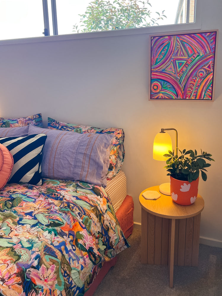 A colorful bedroom corner features a bed with vibrant, patterned bedding. A wooden side table supports a yellow lamp and potted plant. The wall is adorned with My Stores Karma Kaleidoscope Framed Original Painting, while a small window welcomes natural light.