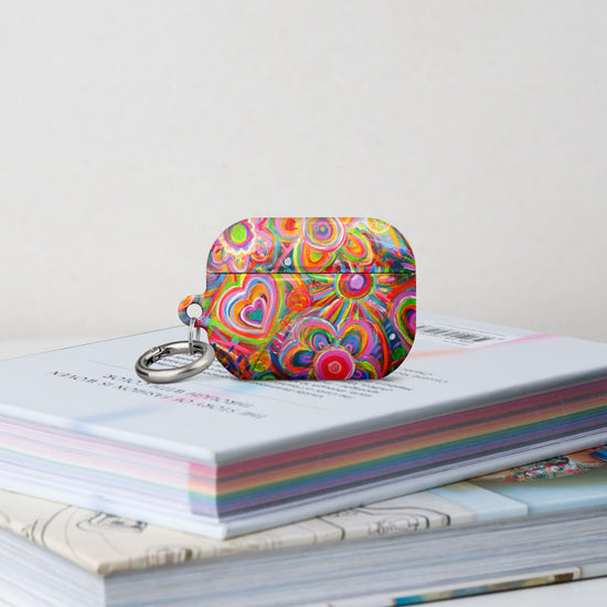 The Love in Neon Airpods Pro Gen 2  Case stands on a stack of books.  It features vibrant heart and floral patterns and swirling rainbow designs. 
