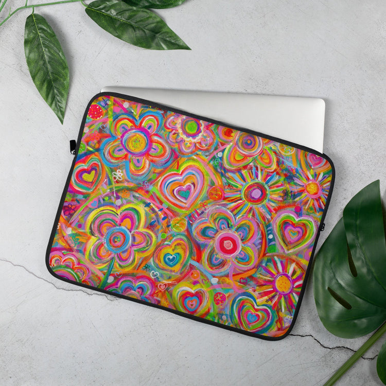 The 15 inch Love in Neon Laptop Sleeve by My Favourite Colour is Rainbow features a vibrant design of multicoloured flowers, hearts and sun. It rests on a grey surface next to green leaves with a laptop partially inside.