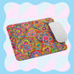 The Love in Neon Mouse Pad by My Favourite Colour is Rainbow features an abstract, vibrant heart and swirl design with a white mouse on top. The background has a wavy blue and pink checkered pattern.