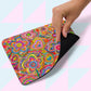 A hand lifts the corner of a Love in Neon mouse pad by My Favourite Colour is Rainbow, featuring vibrant psychedelic heart and floral patterns. The mat has a black underside and lies against a pastel pink and blue geometric background.