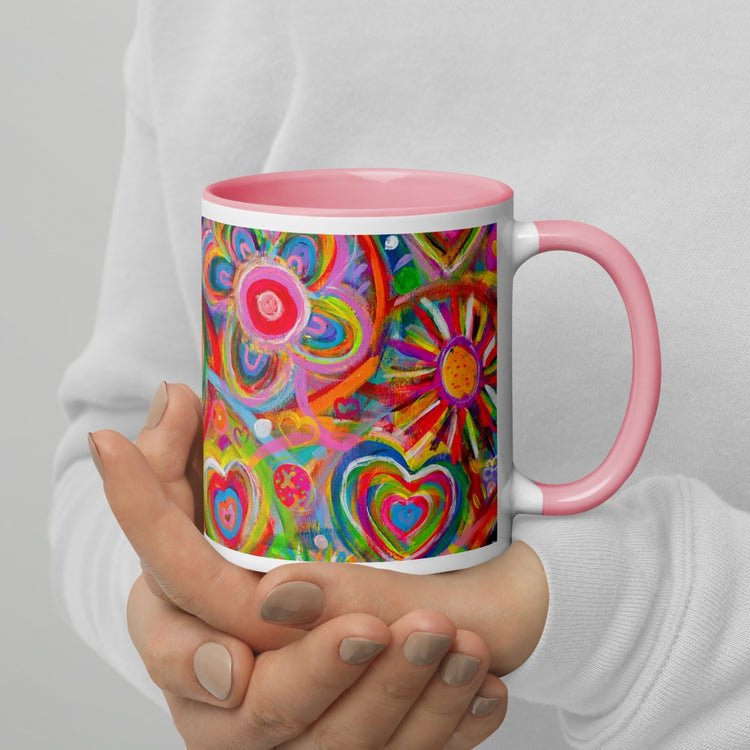 Hands hold the Love in Neon Mug by My Favourite Colour is Rainbow, showing off its vibrant heart, flower, and abstract design. The mugs pink handle and interior contrast with the multicoloured art as the person wears a white top.