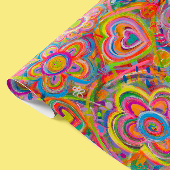 The Love in Neon Wrapping Paper Sheets by My Favourite Colour is Rainbow features vibrant abstract floral and heart patterns, with swirls of pink, yellow, blue, and green.  The slightly folded sheet is shown on a plain yellow background.