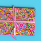 A gift wrapped in My Favourite Colour is Rainbows Love in Neon Wrapping Paper Sheets, with vibrant abstract flowers and hearts.  It has a pink ribbon and is shown against a bright blue background.