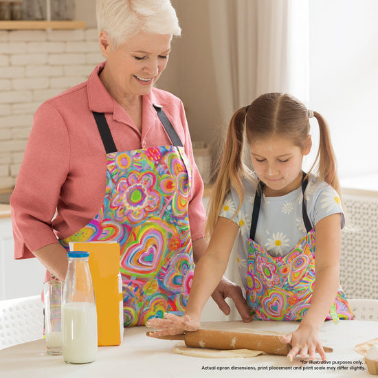 In a kitchen, a smiling older woman and young girl are wearing Love in Neon Aprons.  The girl focuses on rolling dough with a wooden pin. Milk and flour are nearby. The aprons feature vibrant floral and heart designs.