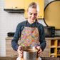 In a kitchen with yellow cabinets, a black oven, and a brick backsplash, someone wearing the Love in Neon Apron by My Favourite Colour is Rainbow holds a cake pan.  The apron features vibrant floral and heart designs.