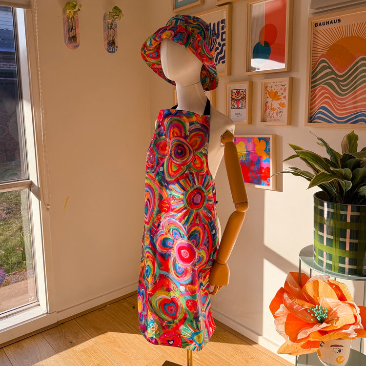 A mannequin displays the Love in Neon Apron by My Favourite Colour is Rainbow, featuring a multicoloured flower and heart design with a matching hat. It stands in a sunlit room with abstract art, an orange paper flower, and a green potted plant, capturing the vibrant cooking experience.