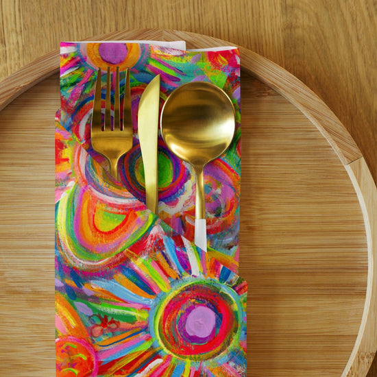 A gold-finished spoon, fork, and knife rest elegantly on a Love in Neon Cloth Napkin by My Favourite Colour is Rainbow, with abstract circular patterns. This chic dining setup sits on a round wooden serving tray.