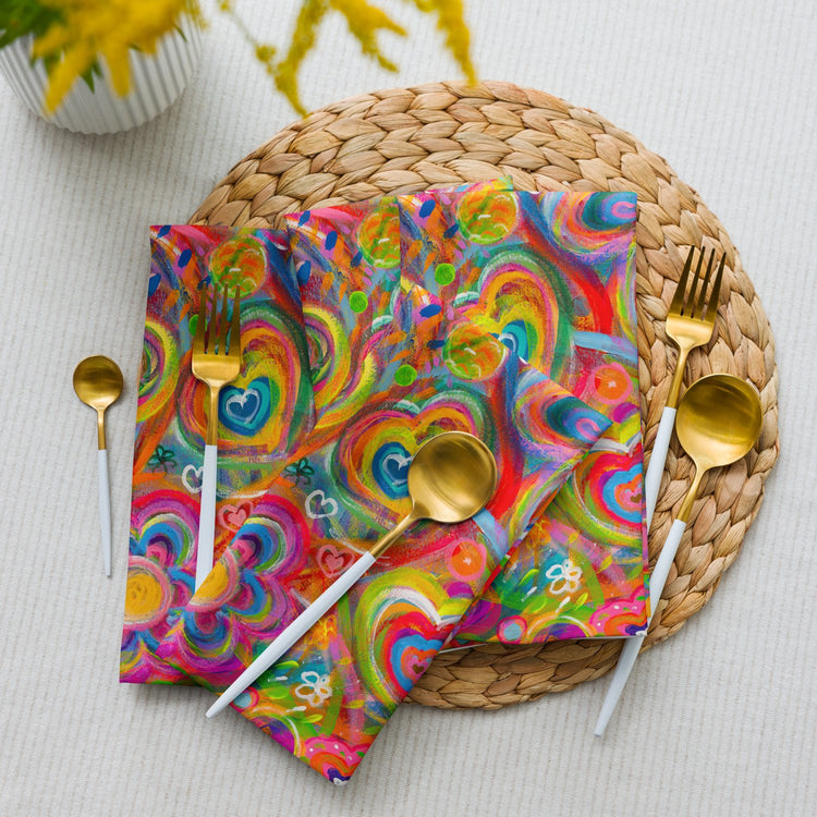 A vibrant table setting features Love in Neon napkins from My Favourite Colour is Rainbow on a round woven placemat. Gold cutlery with white handles enhances the setup, while a white pot of yellow flowers peeks from the corner.