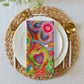 On a white plate sits a napkin from the Mixed Set of 4 Art Napkins by My Favourite Colour is Rainbow, with heart patterns and vibrant colors. The setting includes a woven placemat, gold cutlery, and an abstract art-inspired light-colored tablecloth.