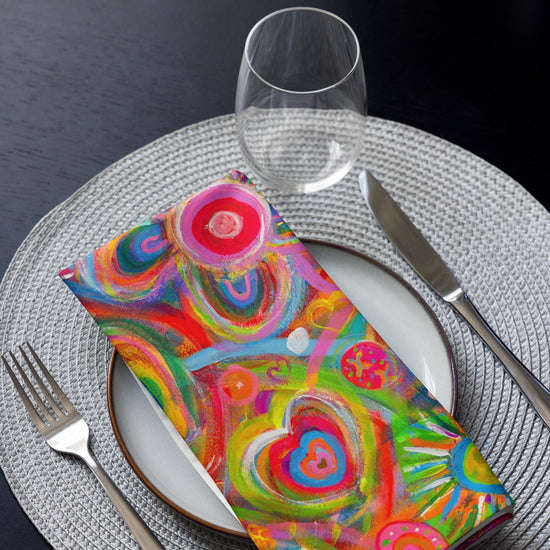 A table setting with a white plate and a vibrant Love in Neon napkin from My Favourite Colour is Rainbow, featuring heart and swirl patterns. A fork, knife, and empty wine glass sit on a grey woven placemat.