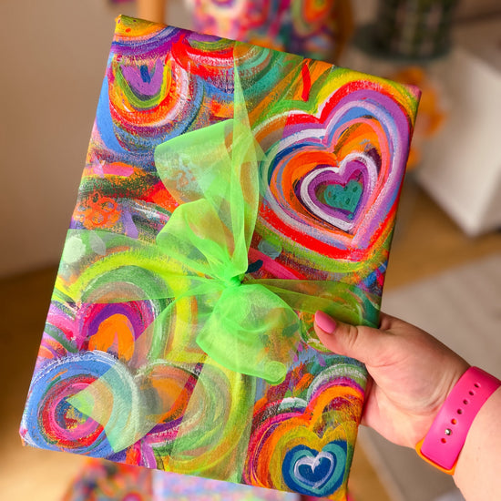 A hand with pink nail polish and a pink watchband holds a gift wrapped in Love in Neon Wrapping Paper Sheets by My Favourite Colour is Rainbow, featuring vibrant abstract rainbow-coloured hearts and flowers, adorned with a green ribbon.