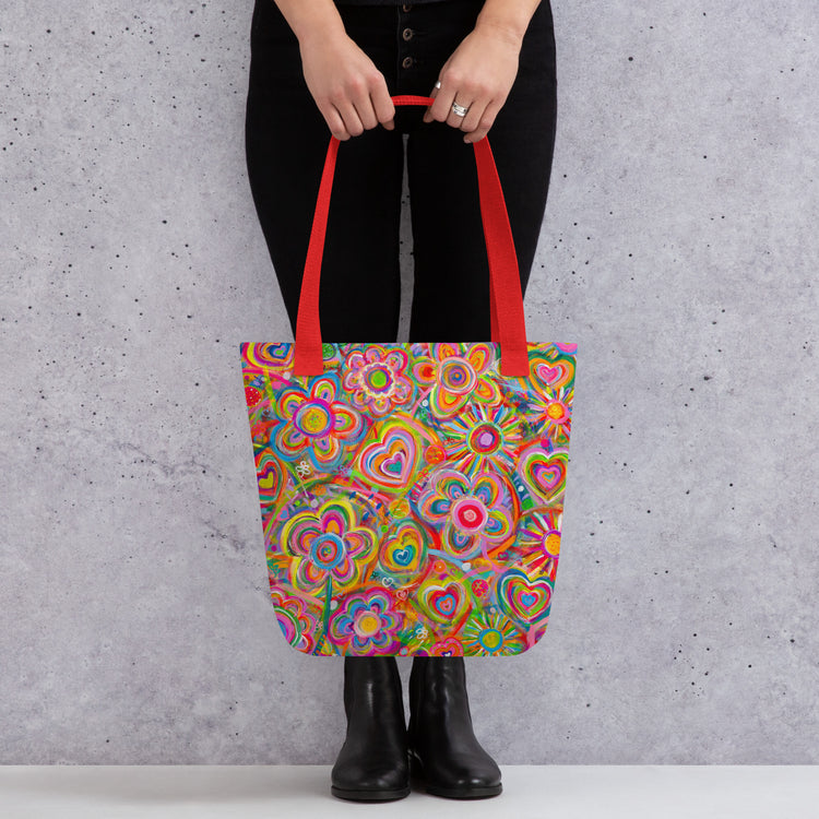 A person holds a Love in Neon Tote Bag by My Favourite Colour is Rainbow with vibrant heart and flower patterns and bright red handles. They stand stylishly against a grey background in black pants and boots.