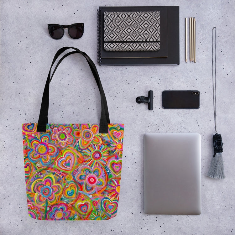 An artful flat lay featuring the Love in Neon Tote Bag by My Favourite Colour is Rainbow, alongside sunglasses, a patterned notebook, a smartphone, a laptop, pens, and a tasseled keychain.