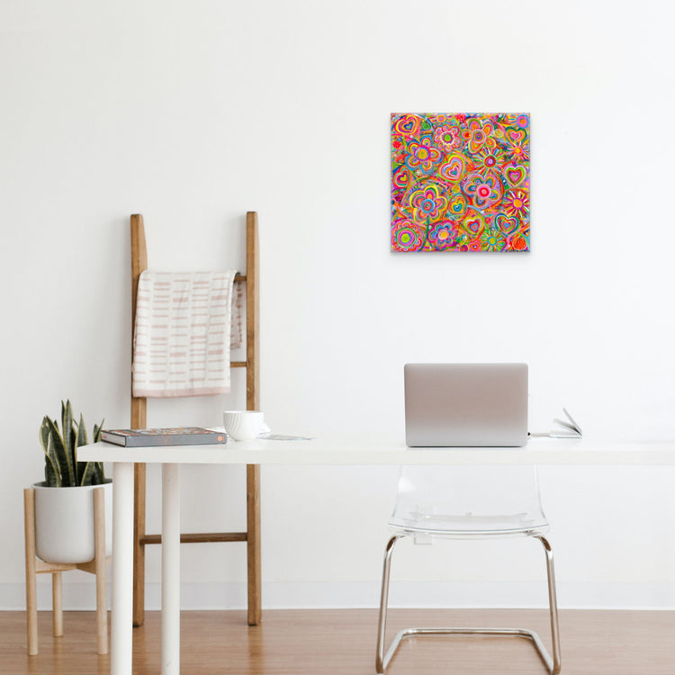 A modern, minimalist workspace features a laptop on a white desk, complemented by a transparent chair and the Love in Neon canvas artwork by My Favourite Colour is Rainbow. A wooden ladder holds a folded blanket, while nearby, a potted plant adds life to the setting.