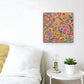 A white room displays a bed with white bedding next to a small round table with a lamp and potted plant. On the wall hangs Love in Neon, vibrant canvas artwork by My Favourite Colour is Rainbow, featuring abstract flowers and hearts.