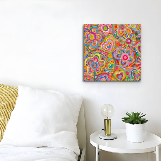 A white room displays a bed with white bedding next to a small round table with a lamp and potted plant. On the wall hangs Love in Neon, vibrant canvas artwork by My Favourite Colour is Rainbow, featuring abstract flowers and hearts.