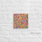 Love in Neon Canvas Artwork by My Favourite Colour is Rainbow features vibrant, abstract art with colorful flowers and hearts on a white textured brick wall. This 12x12 inch piece bursts with pink, orange, and blue swirls.