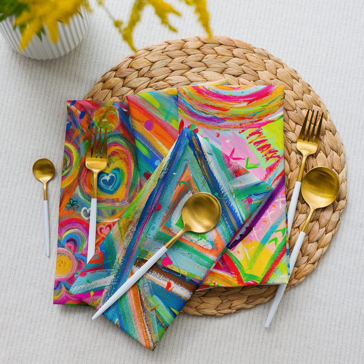 Three gift-wrapped items with abstract designs sit on a woven placemat, accompanied by gold utensils and elegant napkins from the Mixed Set of 4 Art Napkins by My Favourite Colour is Rainbow. A hint of a yellow flower arrangement peeks in, all set against a light fabric background.