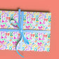 A gift wrapped in My Favourite Colour is Rainbows Multicolour Bow Birthday Wrapping Paper, featuring vibrant patterns of bows, shoes, and hats in pink, blue, orange, and green. It is tied with a light blue ribbon and shown against a coral background.