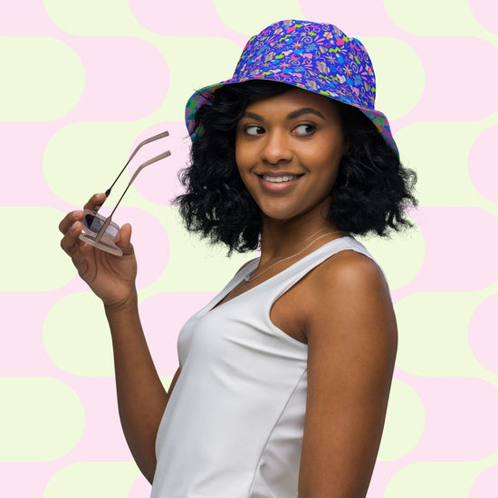 A woman, smiling and holding sunglasses, wears a Neon Carnival/Funfetti Reversible Bucket Hat by My Favourite Colour is Rainbow with a sleeveless white top. The background displays a light green and pink geometric pattern, evoking the vibrant energy of a neon carnival.