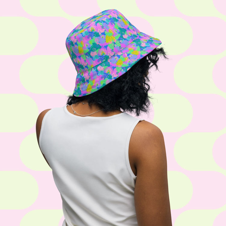 A person with curly black hair is wearing a Neon Carnival/Funfetti Reversible Bucket Hat by My Favourite Colour is Rainbow and a white sleeveless top. They are facing away from the camera, with a backdrop of pastel pink and green circles.