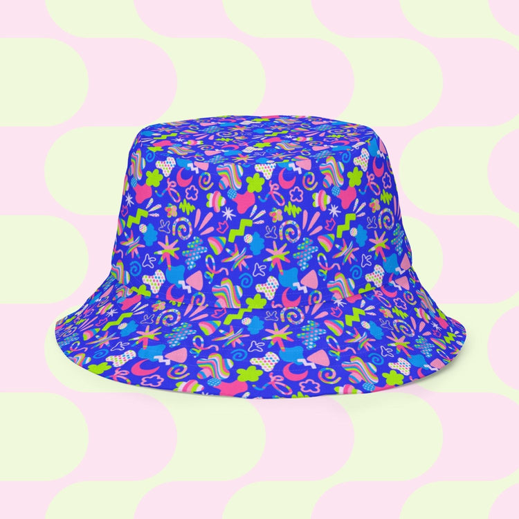 The Neon Carnival/Funfetti Reversible Bucket Hat by My Favourite Colour is Rainbow sits on a pastel backdrop adorned with abstract shapes. It features vibrant doodles, stars, and playful designs in green, pink, yellow, and blue against large soft circles in pink and light green.