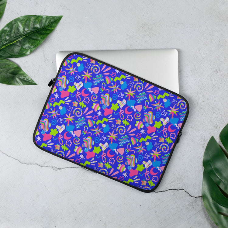 The 13 inch Neon Carnival Laptop Sleeve by My Favourite Colour is Rainbow features a vibrant abstract design in blue, pink, green, and white. Partially open to reveal a laptop inside, its set against a light surface with nearby green leaves.