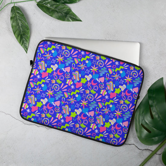 The 15 inch Neon Carnival Laptop Sleeve by My Favourite Colour is Rainbow features a vibrant abstract design in blue, pink, green, and white. Partially open to reveal a laptop inside, its set against a light surface with nearby green leaves.