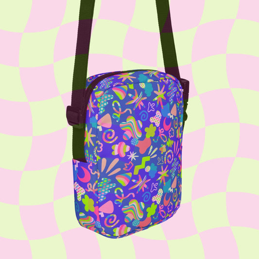 The Neon Carnival Mini Crossbody Bag by My Favourite Colour is Rainbow displays a vibrant, abstract carnival print of flowers, hearts, and stars in pinks, greens, and blues on a purple background. It features adjustable black straps against a wavy pastel green and pink checkered backdrop.