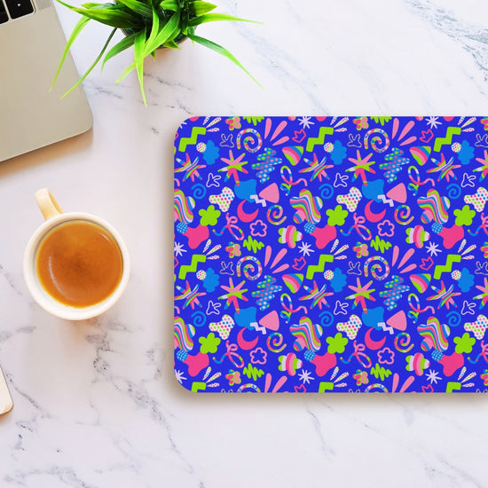 The Neon Carnival Mouse Pad by My Favourite Colour is Rainbow features a vivid abstract pattern of colorful shapes on a blue background, set on a marble surface near a laptop and coffee cup. In the corner sits a small green plant.