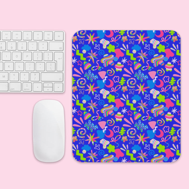 A keyboard and mouse sit on a pink surface beside the Neon Carnival Mouse Pad from My Favourite Colour is Rainbow, showcasing a vibrant abstract design with blue, green, pink, and white shapes.