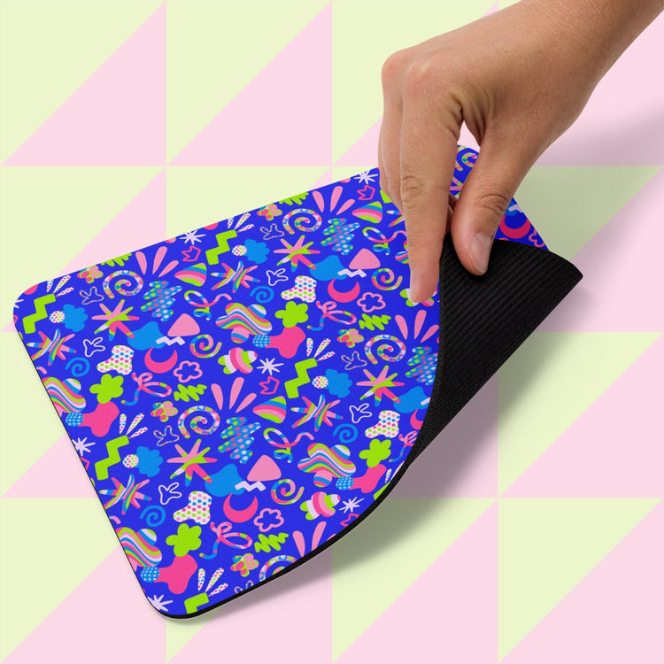 A hand lifts the Neon Carnival Mouse Pad by My Favourite Colour is Rainbow, featuring a vibrant blue background with abstract pink, green, and orange shapes. The underside is black. The backdrop shows a green and pink geometric pattern.