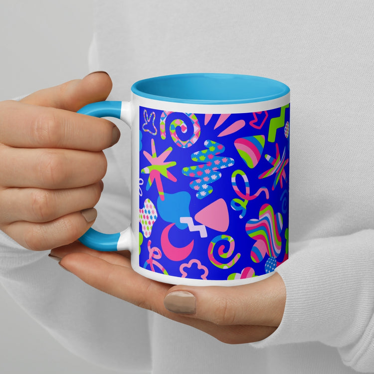 With painted nails, a person holds the Neon Carnival Mug by My Favourite Colour is Rainbow against a blue background. The mugs colourful, abstract design features stars, stripes, various geometric shapes and has a light blue interior.