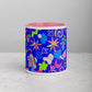 Front view of the Neon Carnival Mug by My Favourite Colour is Rainbow sits on a grey surface, showcasing its vibrant design with abstract pink, green, blue, and white shapes on a bright blue background. Its interior is pink.