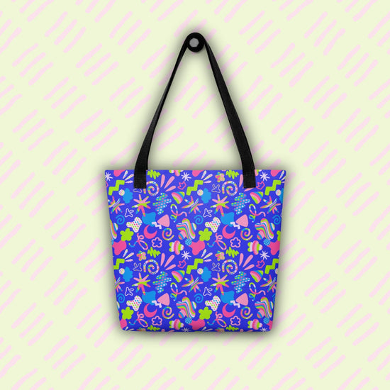 The Neon Carnival Tote Bag by My Favourite Colour is Rainbow with black handles, hangs from a hook. This spacious and colourful tote boasts a vibrant pattern of abstract shapes in blue, green, pink and white.