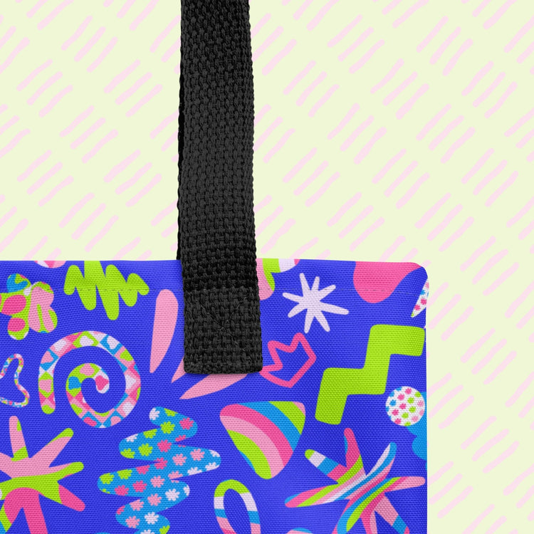 A close-up of the Neon Carnival Tote Bag by My Favourite Colour is Rainbow showing the sturdy black handles. 
 It is a spacious and colourful tote bag which boasts a vibrant pattern of abstract shapes in blue, green, pink and white.  