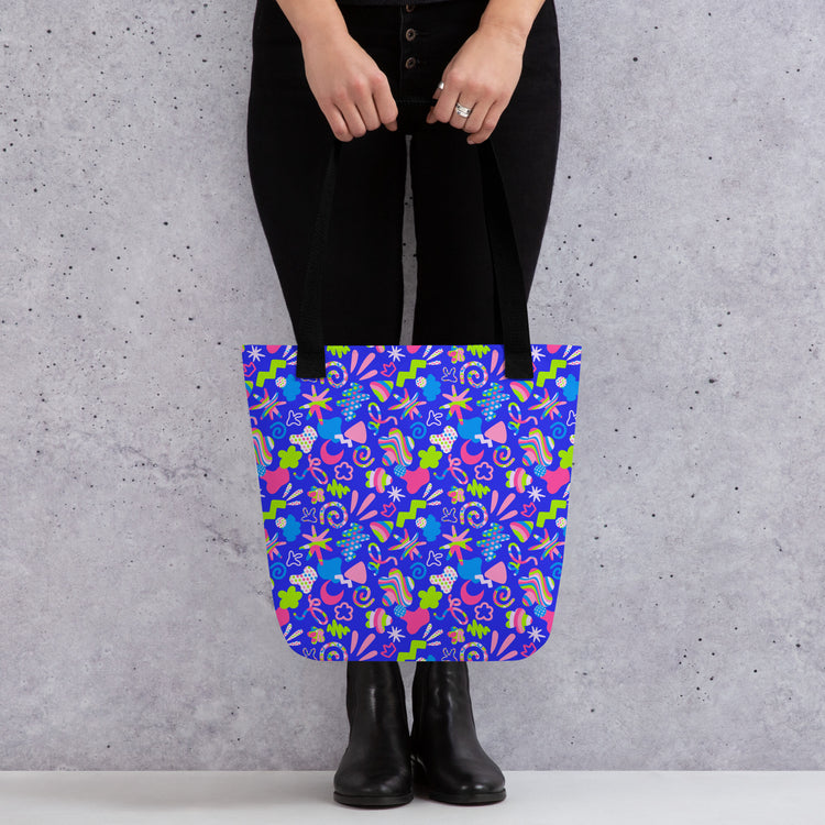 Against a gray textured background, a person in black pants and boots holds the Neon Carnival Tote Bag by My Favourite Colour is Rainbow.  This spacious and colourful tote boasts a vibrant pattern of abstract shapes in blue, green, pink and white.