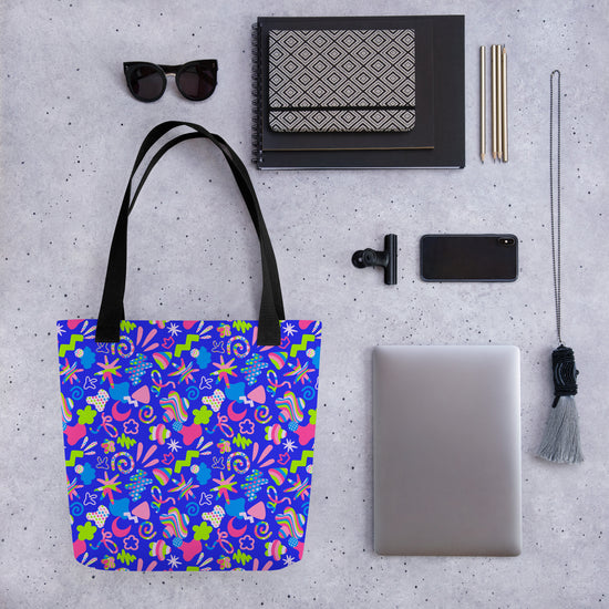 Top-down view of a flat lay on a grey surface featuring a spacious Neon Carnival Tote Bag by My Favourite Colour is Rainbow, sunglasses, notebooks, pens, a smartphone, laptop, small clip, and tassel keychain; all neatly organised. This spacious and colourful tote boasts a vibrant pattern of abstract shapes in blue, green, pink and white.