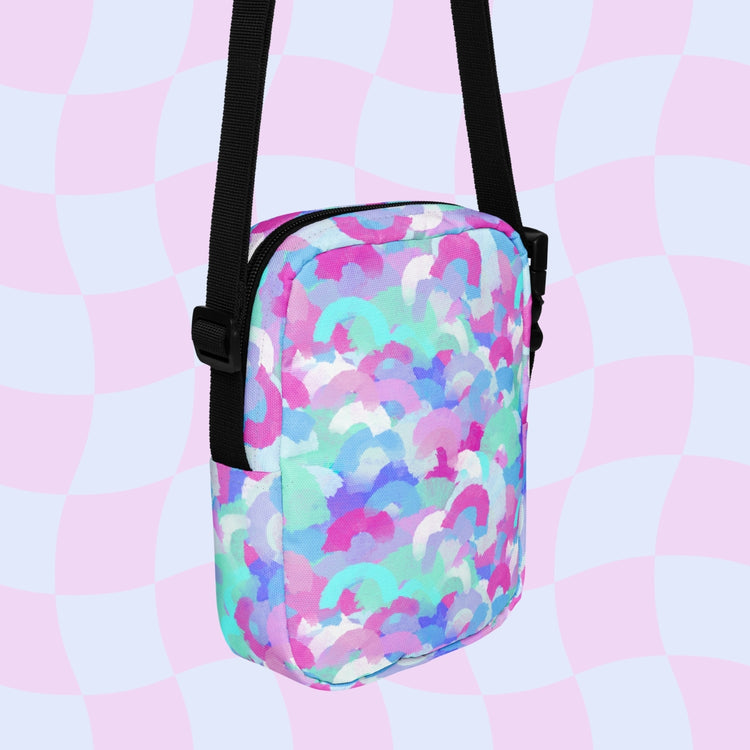 The Pastel Rainbow Mini Crossbody Bag from My Favourite Colour is Rainbow features an abstract pastel rainbow print with pink, purple, blue, and white arcs on a checkered background and includes adjustable black straps.