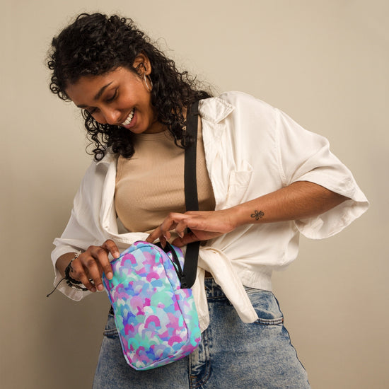 A smiling woman with curly hair stands against a neutral background, holding a Pastel Rainbow Mini Crossbody Bag by My Favourite Colour is Rainbow. She wears a beige top under a white shirt and blue jeans. The bags adjustable straps make it ideal for any outfit.