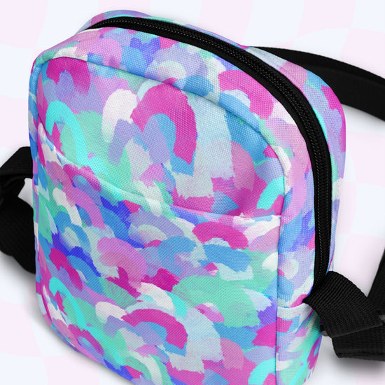 The Pastel Rainbow Mini Crossbody Bag by My Favourite Colour is Rainbow features an abstract pastel design with pink, blue, and purple swirls on a white background. It has a black zipper and adjustable straps.