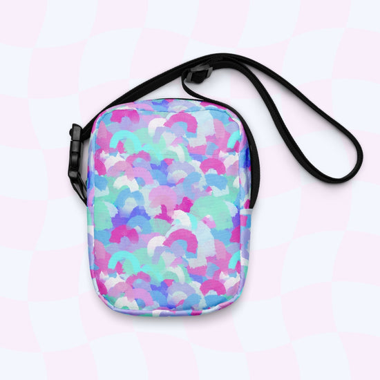 The Pastel Rainbow Mini Crossbody Bag by My Favourite Colour is Rainbow is displayed against a light, checkered background. It features adjustable straps and a vibrant mosaic of overlapping pastel blue, pink, and purple shapes reminiscent of a rainbow print.