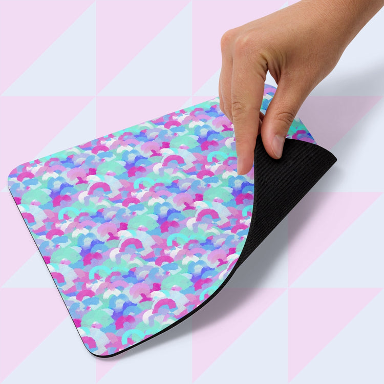 A hand lifts the corner of the Pastel Rainbow Mouse Pad by My Favourite Colour is Rainbow, featuring a vibrant abstract pattern in pink, blue, purple, and white on top with a black underside. The background displays a geometric pastel pattern.