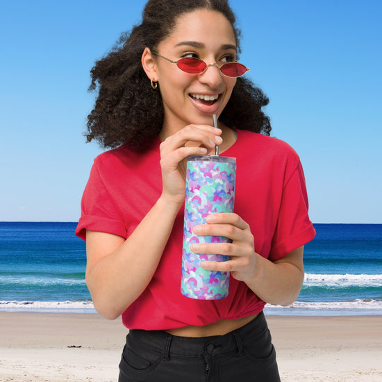 A person in a red shirt and sunglasses drinks from a My Favourite Colour is Rainbow Pastel Rainbow Stainless Steel Tumbler with a metal straw, standing on the beach with curly hair tied back against the backdrop of clear skies and ocean waves.