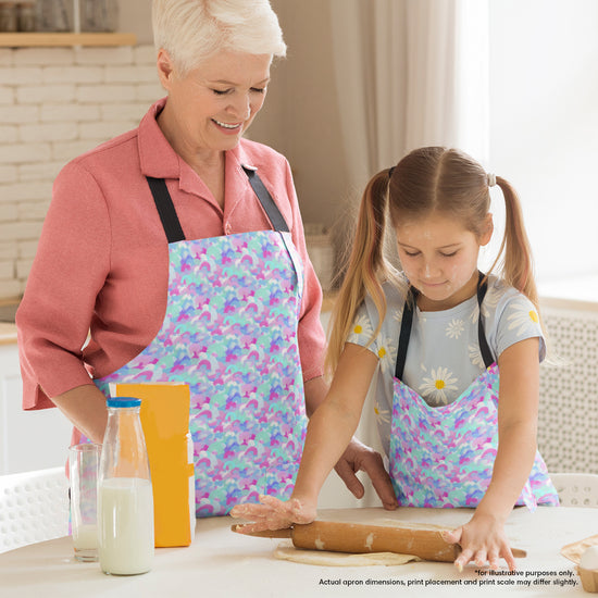 A woman and young girl happily wear matching Pastel Rainbow Aprons by My Favourite Colour is Rainbow while making dough. The counter holds a rolling pin, milk and  flour in their bright kitchen.  The aprons feature a soft pastel pattern of blue, pink, and white rainbow shapes.